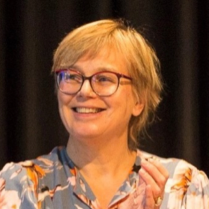 Eva Maria Welskop-Deffaa hat kurze blonde Haare und trägt eine rote Brille zur gemusterten Bluse. Sie strahlt freudig, schaut dabei aber nicht in die Kamera.