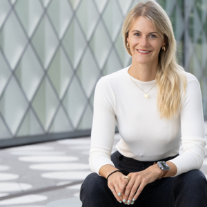 Zarah Bruhn hat lange blonde Haare und trägt eine dunkle Anzughose und einen weißen Pullover und lächelt in die Kamera.